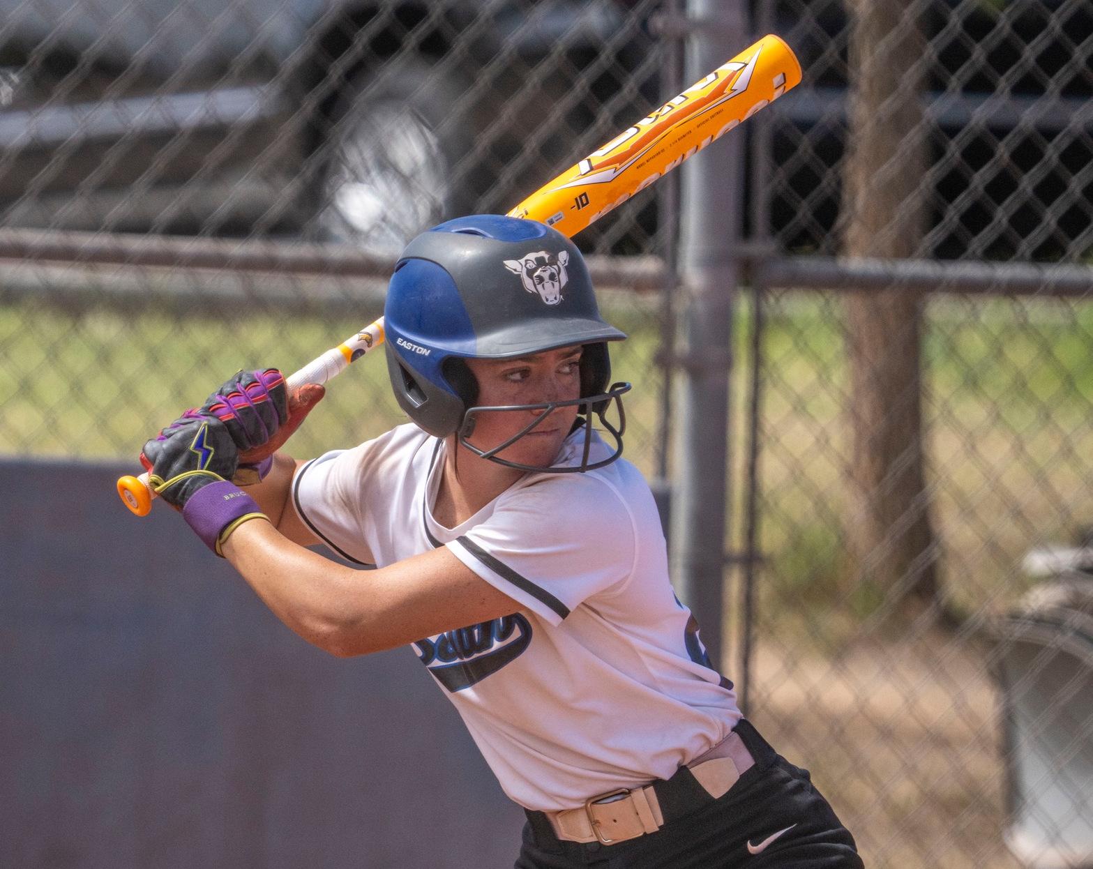 Photo: Softball player in a white and black uniform preparing to bat during a game. The athlete is wearing a black helmet and colorful batting gloves while holding an orange bat over their shoulder in the batter&rsquo;s box. A chain-link fence and blurred background are visible behind the player.

