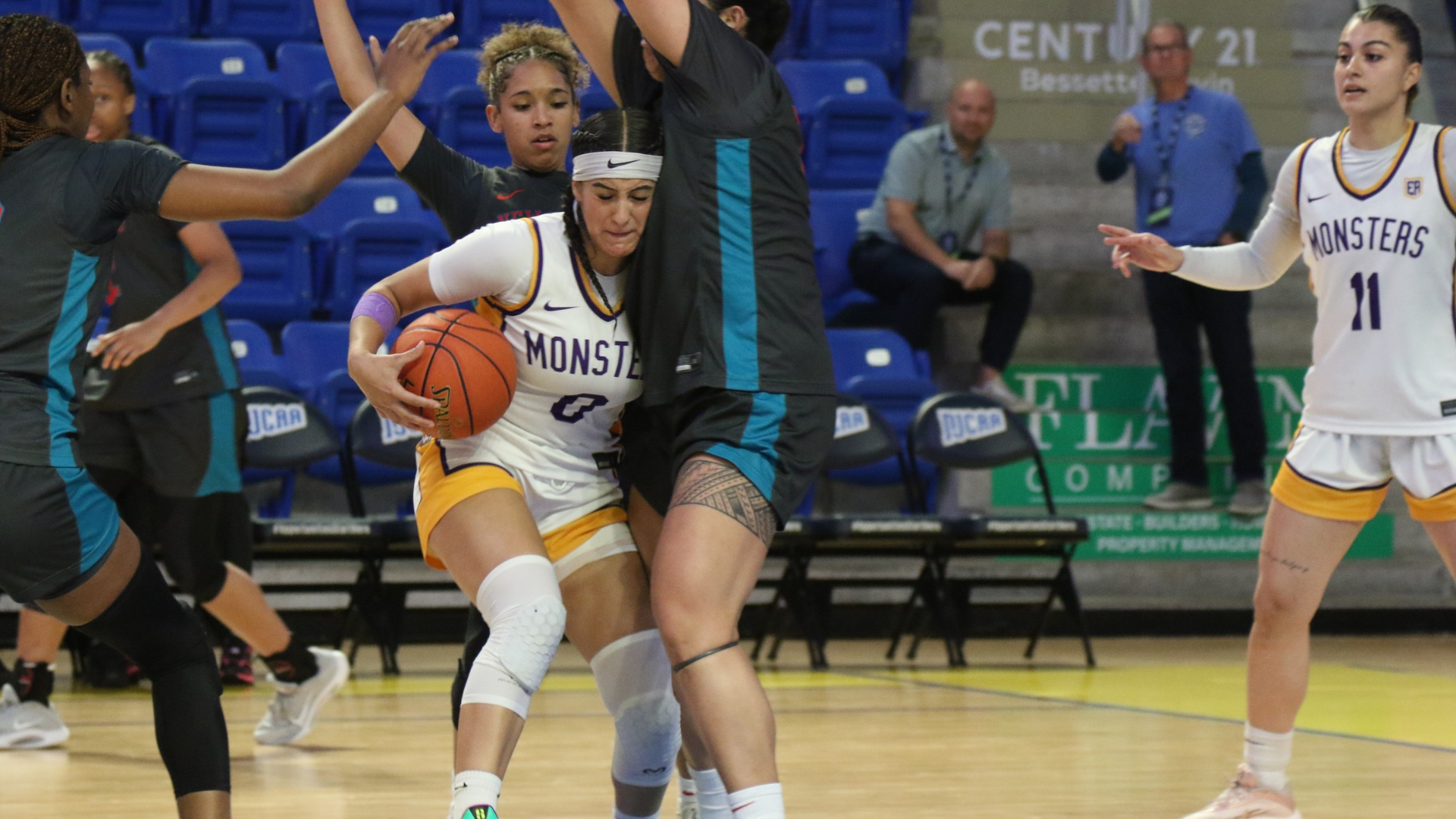 Photo: Esmeralda Enriquez dribbles through traffic during a game, driving toward the basket while being closely defended by multiple opponents. She wears a white &ldquo;Monsters&rdquo; jersey with a headband and knee sleeve, protecting the ball as defenders raise their arms to contest. A teammate stands to the right ready for a pass, with the bench and spectators visible in the background.
