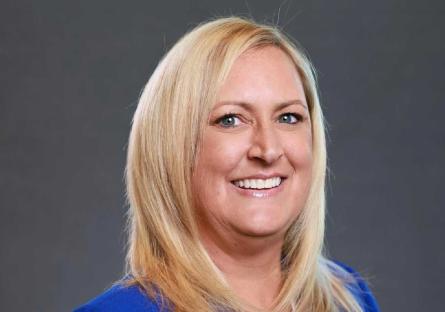 Photo: Headshot of Christina Hundley smiling against a dark gray background. She has shoulder-length blonde hair and is wearing a royal blue top.
