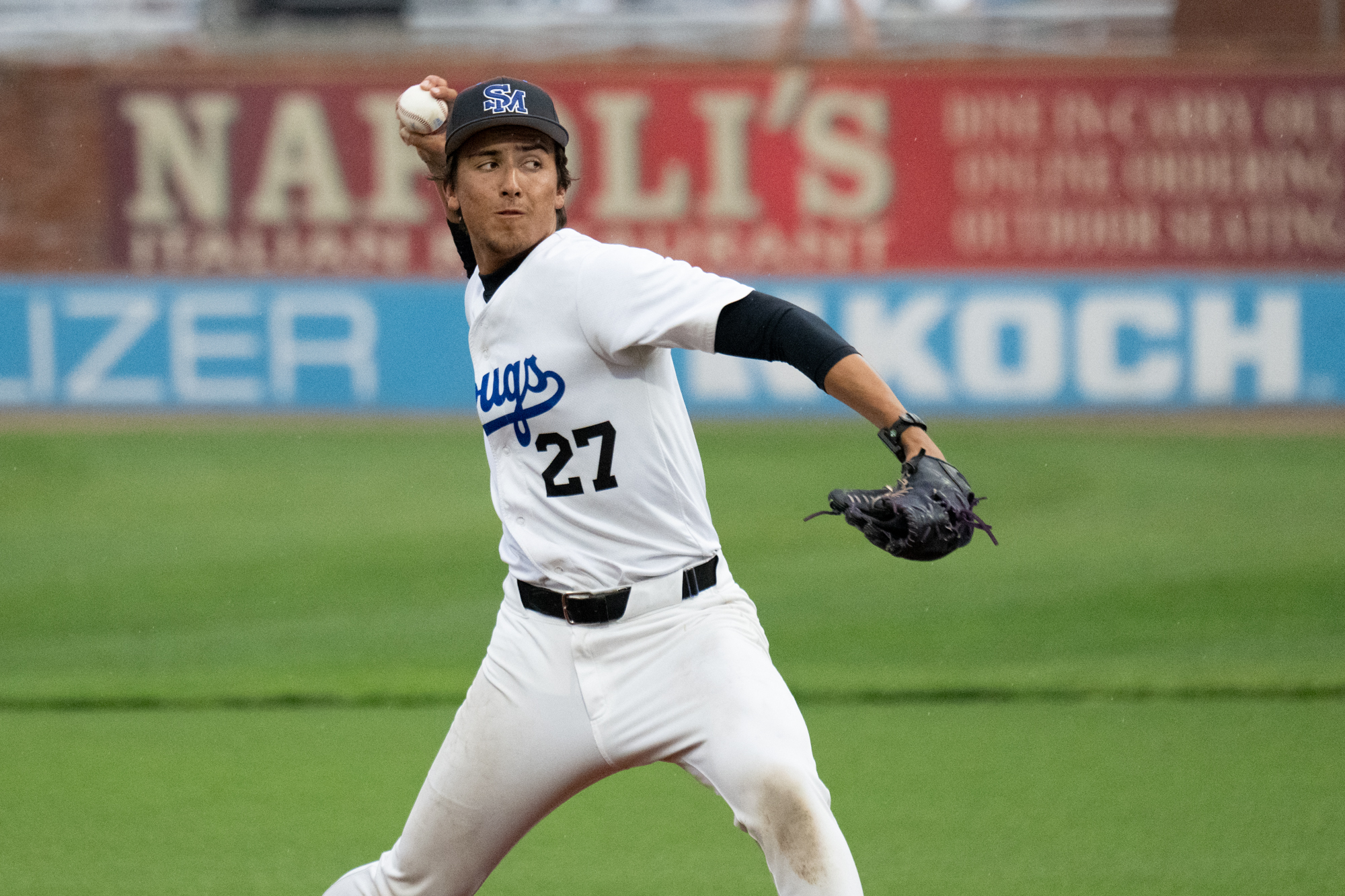 Photo: Baseball player in a white South Mountain Community College uniform prepares to throw a pitch during a game, wearing jersey number 27 and a blue cap while standing on the mound with a blurred outfield advertisement in the background.